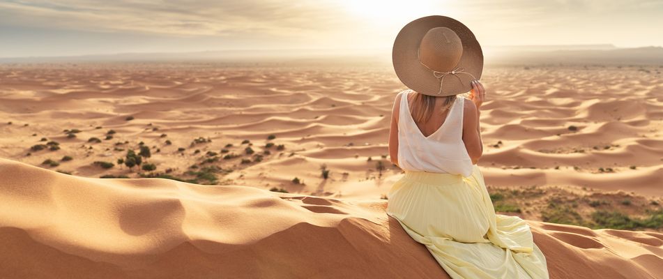 Frau mit Sonnenhut sitzt in der Sahara in Marokko und blickt auf die weite Landschaft