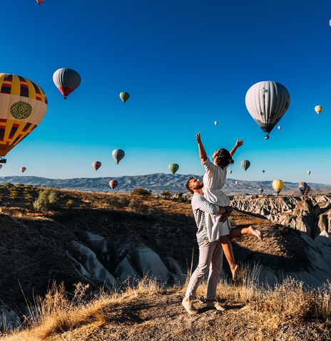 Frau mit ausgebreiteten Armen wird von Mann hochgehoben während sie die Heißluftballons über den Feenkaminen in Kappadokien in der Türkei bewundern