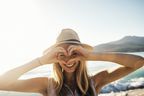 Frau mit Sommerhut am Strand formt mit den Händen ein Herz und schaut hindurch