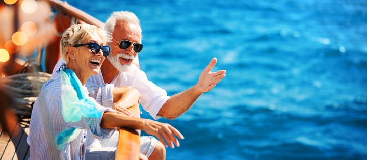 Best-Ager-Paar sitzt an der Reling eines Boots, trägt Sonnenbrillen und hält die Füße über dem Wasser