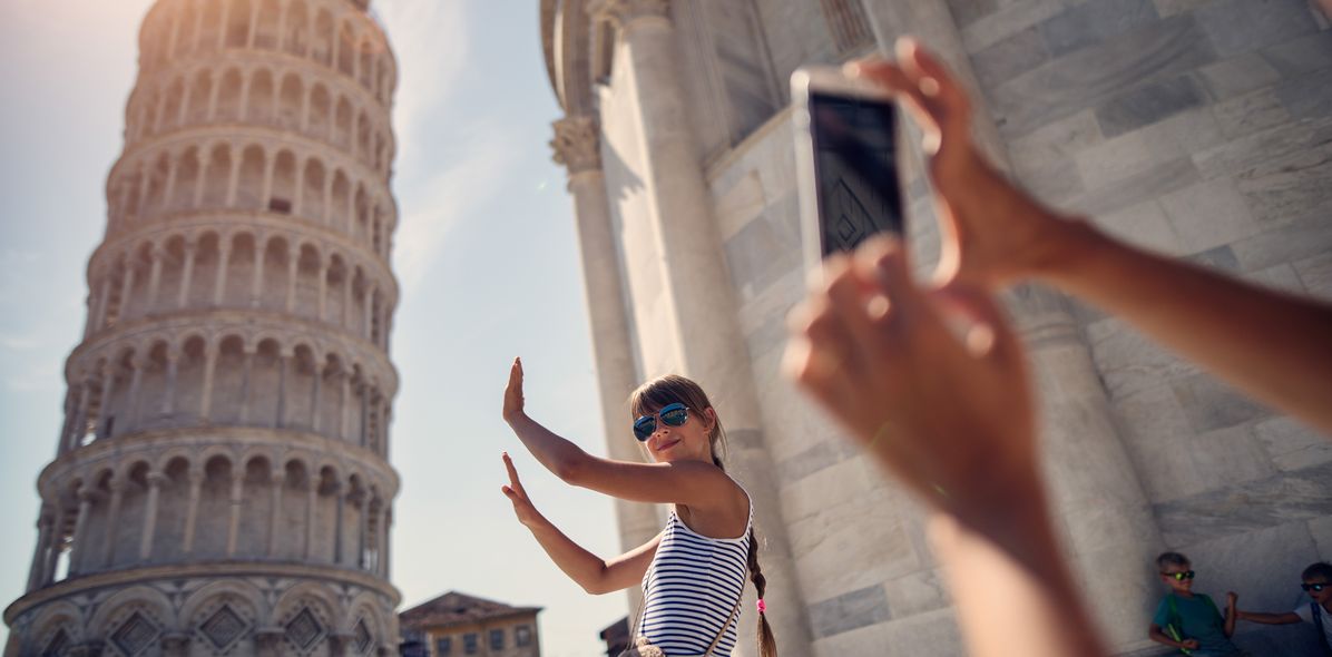 Junges Mädchen steht mit der typischen Handpose vor dem schiefen Turm von Pisa und wird mit einem Handy fotografiert