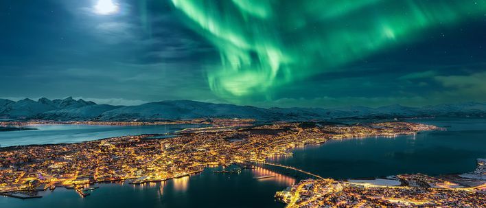 Blick auf die verschneite Stadt Tromso von oben bei Nacht und Nordlichter am Himmel, Norwegen