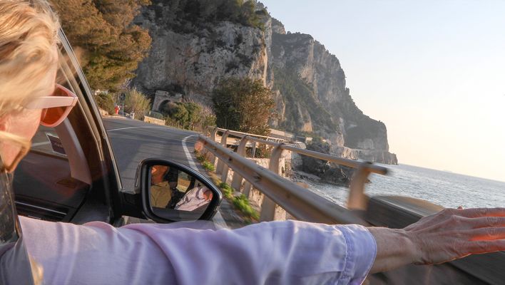 Eine Frau streckt auf einer Küstenstraße die Arme aus dem Cabrio und blickt auf Felsen und Meer.