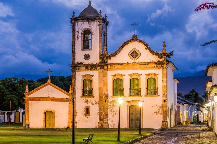 Kirche Santa Rita in der historischen Altstadt von Paraty, Brasilien, mit Kopfsteinpflastergasse und beleuchteten Straßenlaternen in der Abenddämmerung