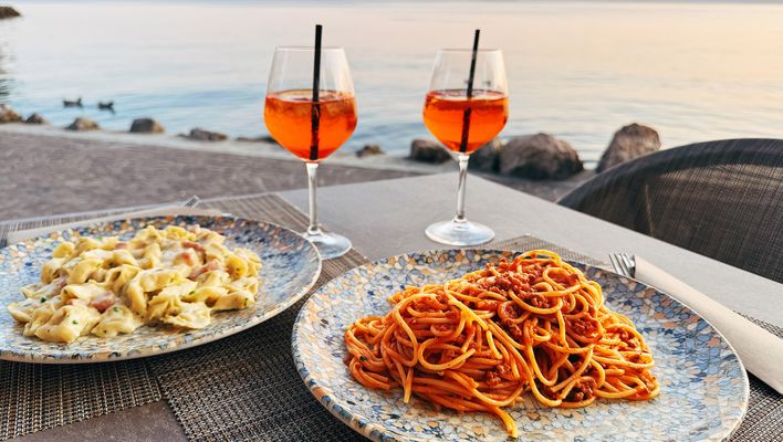 Teller mit Pasta und Gläser Aperol Spritz auf einem Tisch mit Blick auf den Gardasee bei Sonnenuntergang