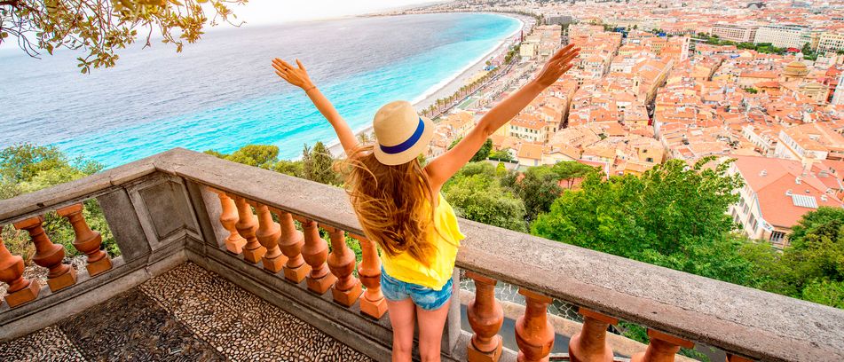 Frau mit Hut und ausgestreckten Armen blickt von einer Aussichtsterrasse über die Küste und Stadt von Nizza