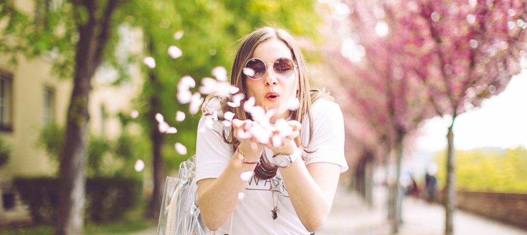 Frau pustet Kirschblüten in die Luft, blühende Bäume im Hintergrund, Frühlingsatmosphäre
