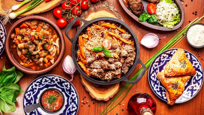Verschiedene usbekische Gerichte mit Reis, Fleisch, Gemüse und Brot stehen in bunt verzierten Schalen bereit