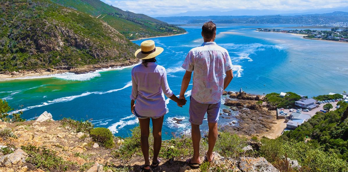 Ein Paar, das händchenhaltend auf einer Klippe in Knysna auf der Garden Route in Südafrika aufs weite Meer blickt