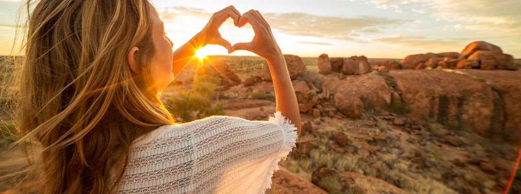 Eine Frau formt mit ihren Händen ein Herz über dem Sonnenuntergang im Outback von Australien