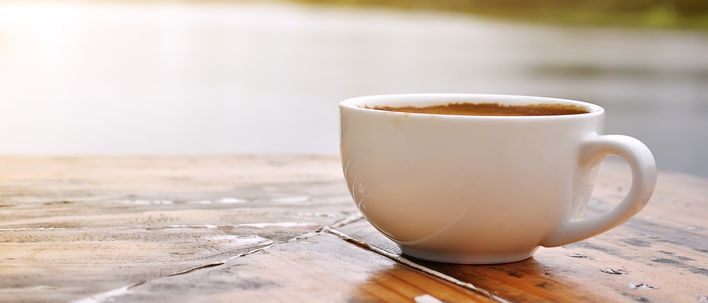 Weiße Kaffeetasse auf einem Holztisch mit Blick auf Wasser im warmen Morgenlicht