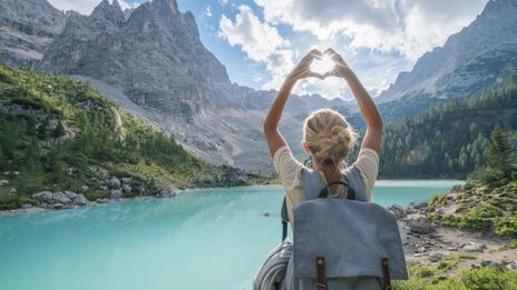 Frau von hinten fotografiert, steht vor einer weitläufigen Seenlandschaft in Italien und fängt mit ihrer zum Herz geformten Hand die Sonne ein