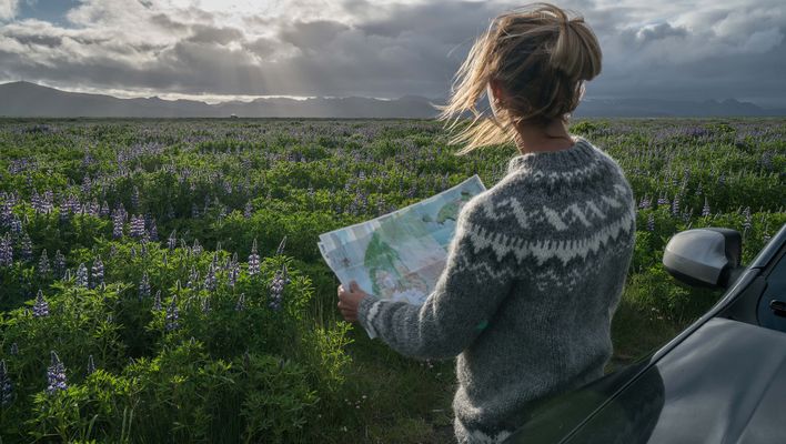 Frau mit einer Landkarte in der Hand lehnt an ihrem Auto und blickt auf eine Landschaft Islands