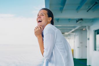 Eine Frau steht lächelnd auf dem Deck eines Kreuzfahrtschiffs und blickt über das Meer.