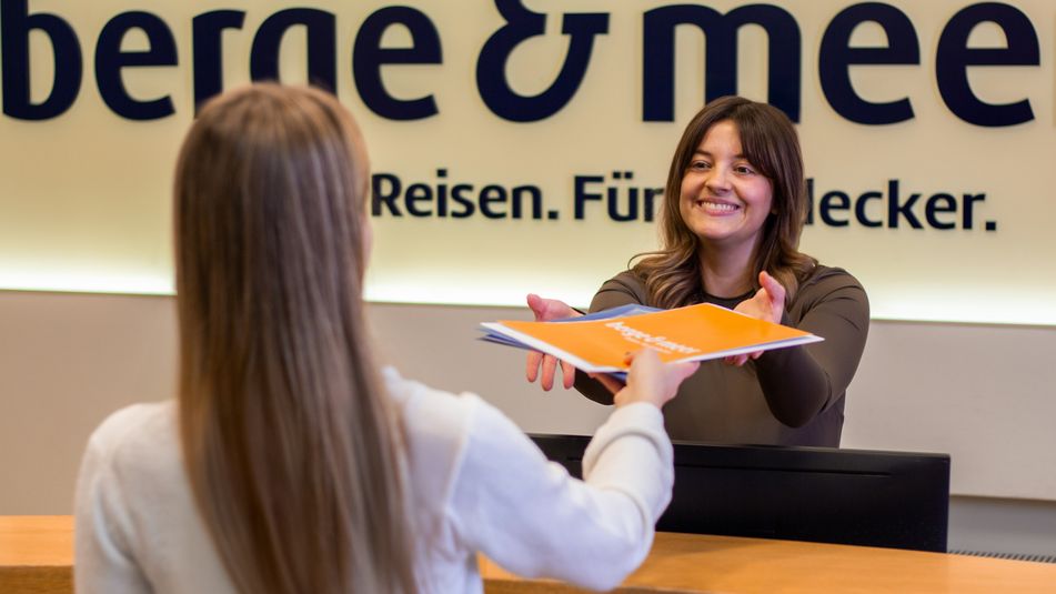 Eine Person überreicht einer Kollegin Ihre Unterlagen am Empfang von Berge & Meer