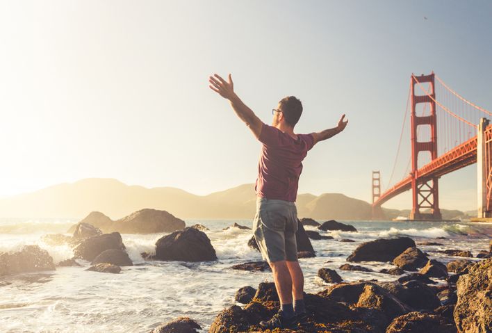 Mann mit ausgebreiteten Armen steht neben der Golden Gate Bridge in San Francisco und schaut Richtung Wasser