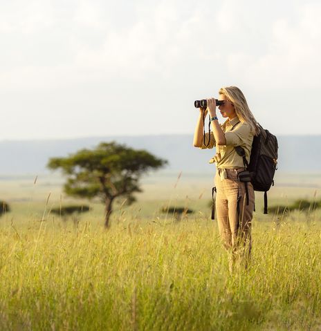 Frau steht während einer Safari mit Fernglas in weiter Graslandschaft mit Akazienbäumen im Hintergrund 