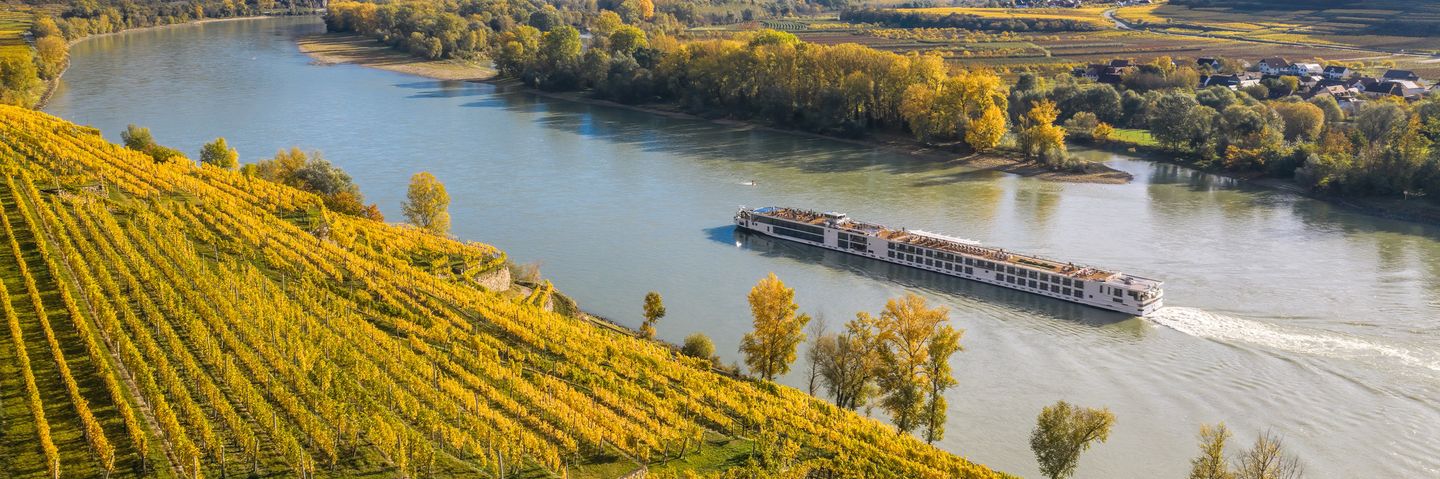 Flusskreuzfahrtschiff fährt entlang der Donau in Österreich mit Weinbergen am Ufer