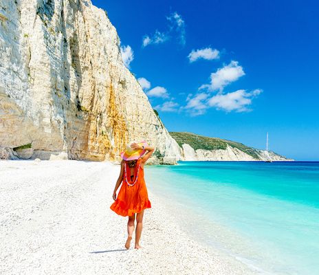 Frau von hinten spaziert auf weißem Sandstrand von Argostoli am türkisblauem Meer mit Felsen umrahmt in Kefalonia in Griechenland