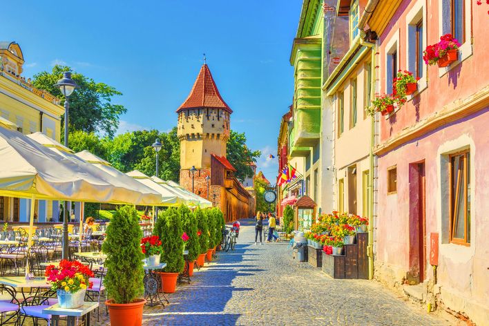 Eine gepflasterte Gasse in Sibiu führt zwischen bunten Häusern und Blumen zu einem alten Stadtturm.
