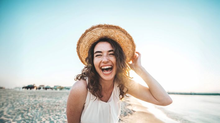 Frau mit Sonnenhut steht am Strand und lacht in die Kamera