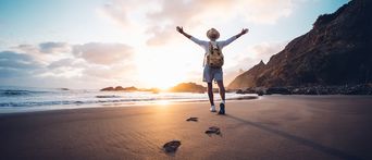 Reisender mit Rucksack genießt den Sonnenuntergang am Strand mit ausgebreiteten Armen