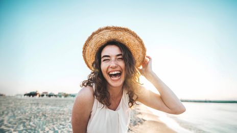 Frau mit Sonnenhut steht am Strand und lacht in die Kamera