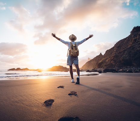 Reisender mit Rucksack genießt den Sonnenuntergang am Strand mit ausgebreiteten Armen