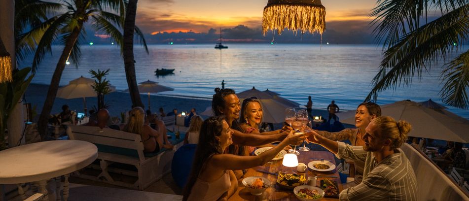 Gruppe junger Leute sitzt beim romantischen Abendessen am Strand unter Palmen