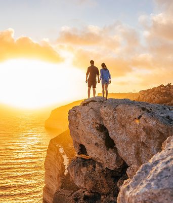 Zwei Personen stehen auf Felsen und schauen auf das Meer beim Sonnenuntergang auf Malta