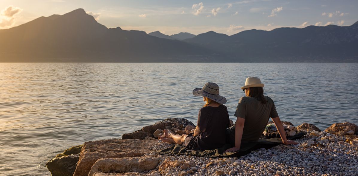 Paar sitzt am Ufer des Gardasees und genießt den Sonnenuntergang mit Blick auf die Berge