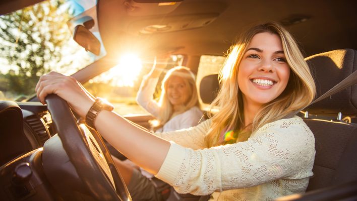 Zwei Frauen sitzen in einem Auto und lächeln in die Kamera während die Sonne ins Auto scheint