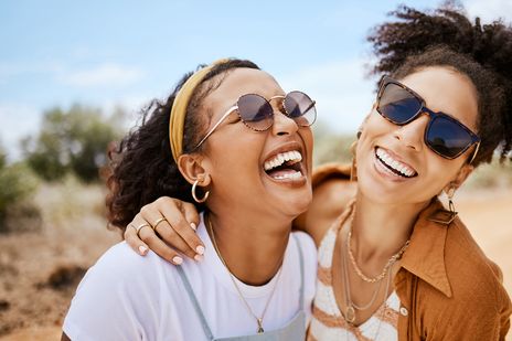 Zwei Freundinnen lachen gemeinsam in der Sonne und tragen Sonnenbrillen