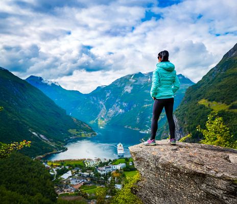 Eine Frau steht auf einem Felsvorsprung und schaut in einen Fjord hinunter wo ein Schiff zu sehen ist