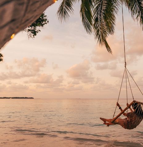 Frau in Hängeschaukel unter Palmen am Strand mit Blick aufs Meer im Abendlicht