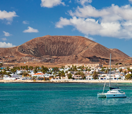 Ein Segelboot liegt vor einer Küstenstadt auf Fuerteventura, hinter der ein kahler Vulkanberg aufragt