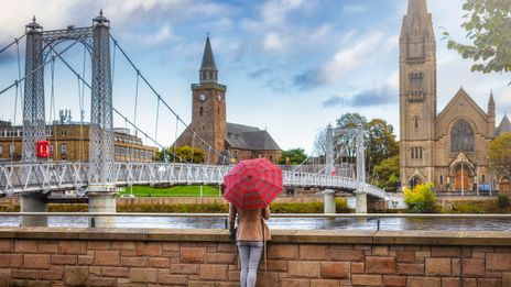 Frau mit rotem Regenschirm steht am Fluss Ness mit Blick auf die Brücke der Stadt Inverness