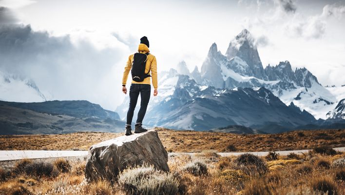 Mann mit Rucksack in Rückansicht steht auf einem Felsstück und blickt auf die Berge von El Chaltén im argentinischen Patagonien