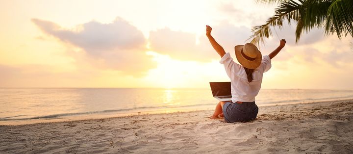 Frau sitzt mit Laptop am Strand vor dem Meer und genießt den Sonnenaufgang