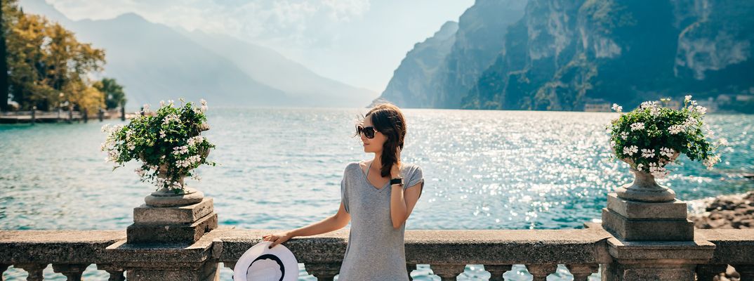 Frau in Sommerkleid und Hut steht an einer Promenade mit Blick auf den Gardasee