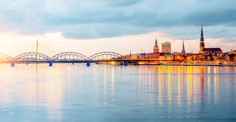 Riga bei Abendlicht mit beleuchteter Brücke, Fluss und moderner Skyline