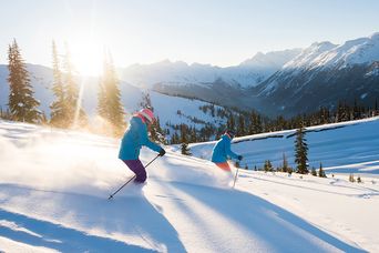 Zwei Skifahrer bei der Abfahrt auf schneebedeckter Piste mit Bergen und Wald im Hintergrund