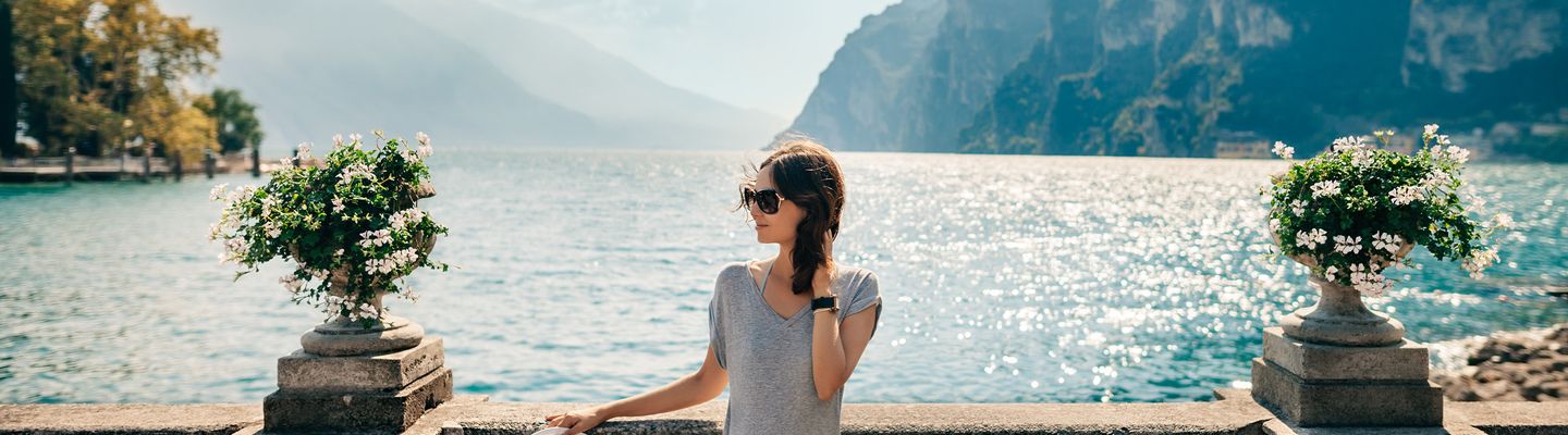 Frau in Sommerkleid und Hut steht an einer Promenade mit Blick auf den Gardasee