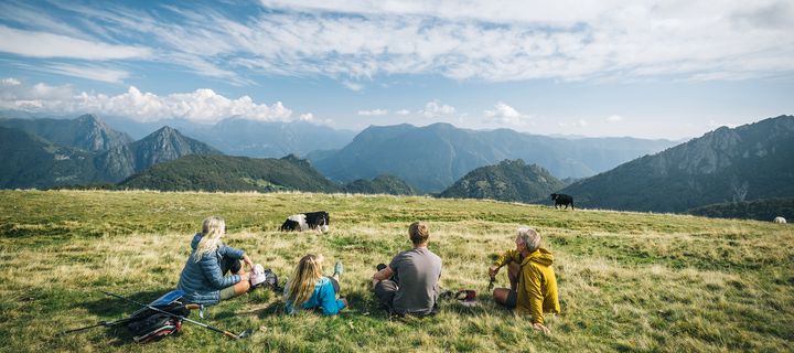Wandergruppe genießt Rast auf grüner Almwiese mit Bergpanorama im Hintergrund