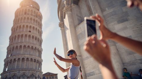 Junges Mädchen steht mit der typischen Handpose vor dem schiefen Turm von Pisa und wird mit einem Handy fotografiert