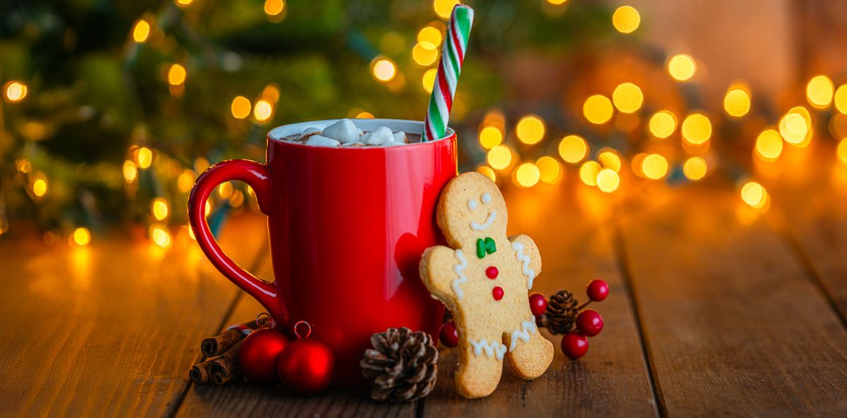 Ein Lebkuchenmännchen steht vor einer roten Weihnachtstasse mit Kakao vor einem beleuchtetem Weihnachtsbaum mit Berge&Meer Superzeichen