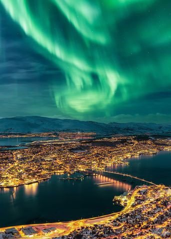 Blick auf die verschneite Stadt Tromso von oben bei Nacht und Nordlichter am Himmel, Norwegen