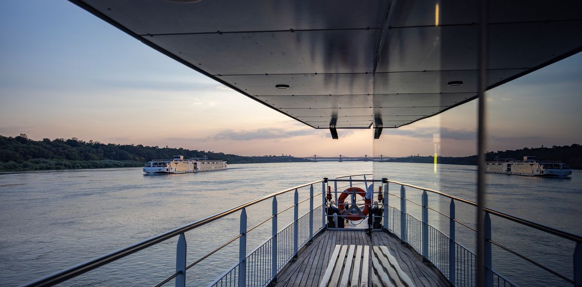 Blick auf das Außendeck eines Flusskreuzfahrtschiffes bei Sonnenuntergang mit Flusslandschaft im Hintergrund