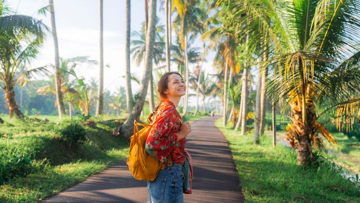Reisende Frau mit Rucksack auf einem Weg zwischen Palmen in tropischer Landschaft auf Bali
