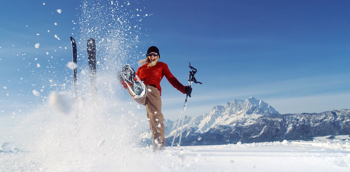 Skifahrerin vor verschneiter Berglandschaft wirbelt Schnee mit ihrem Fuß auf, während ihre Skier neben ihr im Schnee stecken